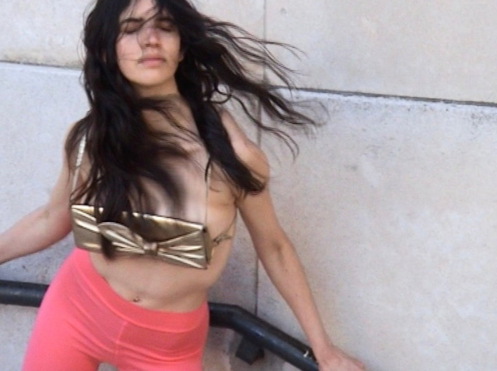 Woman wearing a gold bag-top and pink leggings against a light-colored wall.