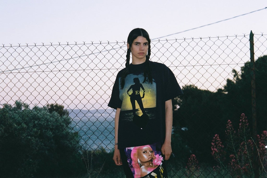 Person wearing a graphic t-shirt with a female silhouette design, standing in front of a chain-link fence.