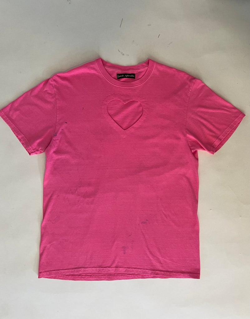 A bright pink t-shirt with a heart cut-out in the center, displayed flat on the floor