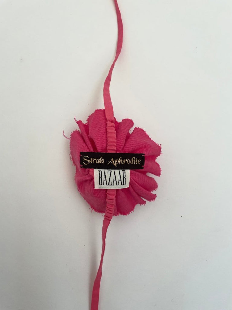 Pink fluffy flower accessory with frayed edges and elastic ties and a Sarah Aphrodite black label, displayed against a white background