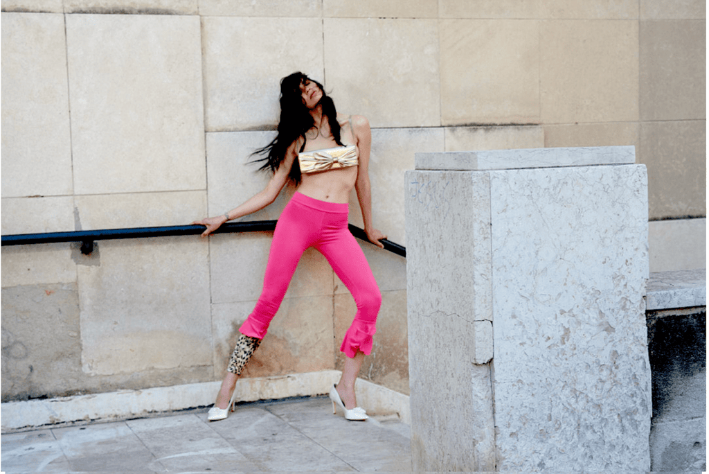 Woman in pink leggings and gold top and cheetah leg piece leaning against a wall.