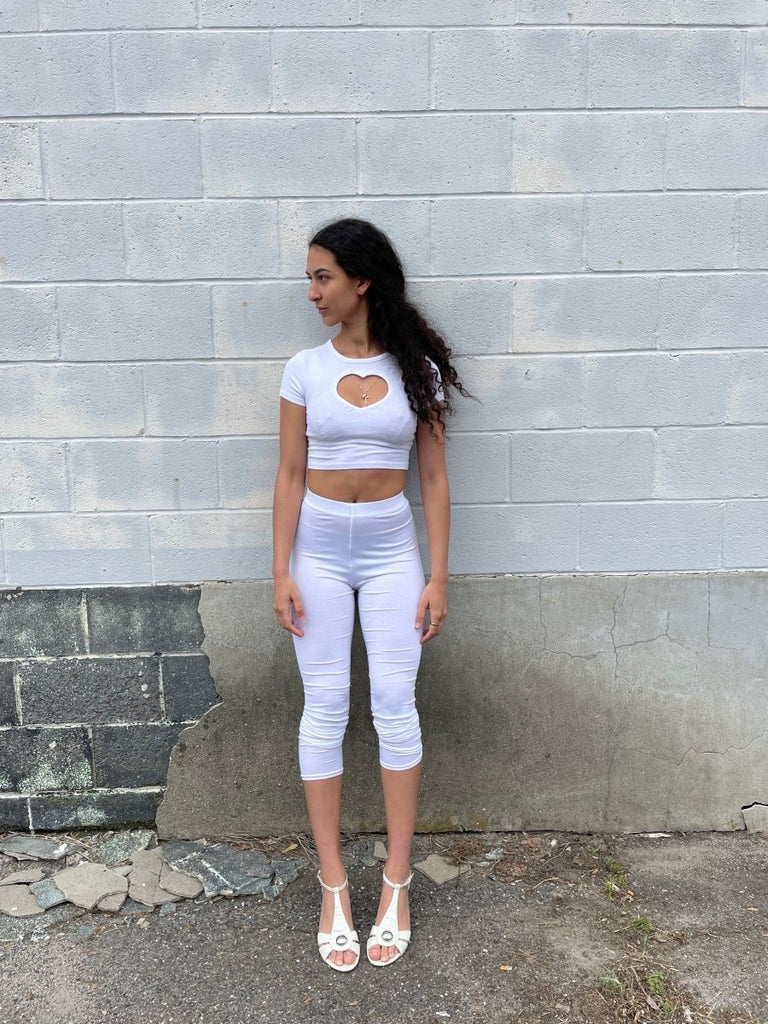 Person wearing a white crop top with a heart cutout and white pants against a white brick wall.