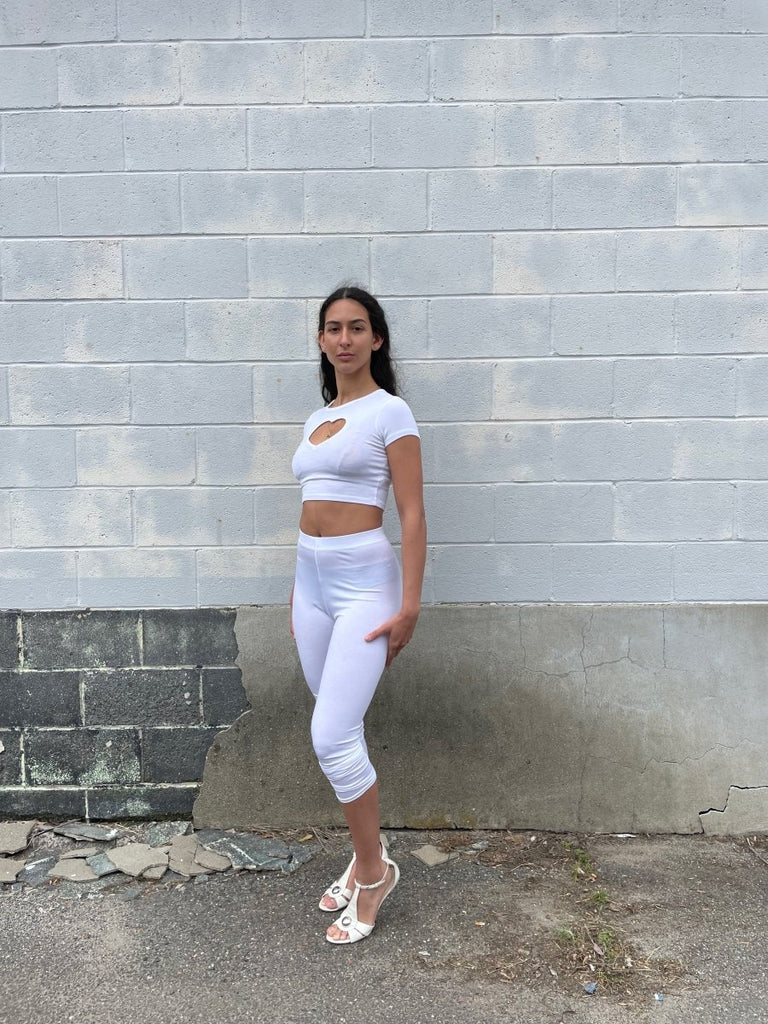 Person wearing a white crop top with a heart cutout and white pants against a white brick wall.