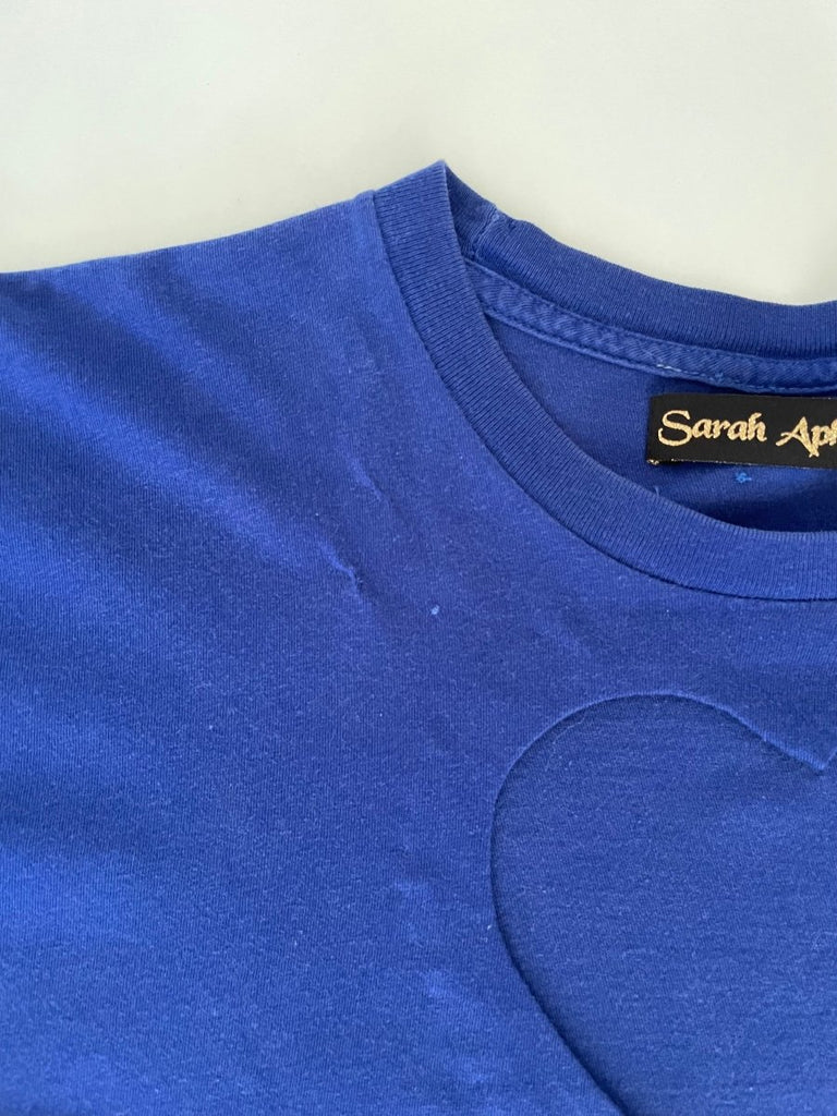 detail of a mended hole in an Ultramarine blue t-shirt with a heart cut-out design in the center, displayed flat on the floor