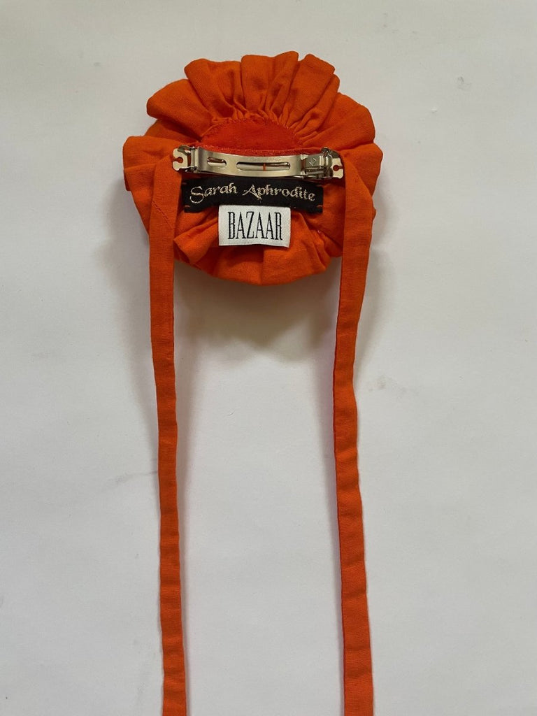 Handmade one-of-a-kind orange flower hair clip by Sarah Aphrodite, featuring a purple button gem center, silver clip, and long ties made from upcycled SA scrap fabrics.